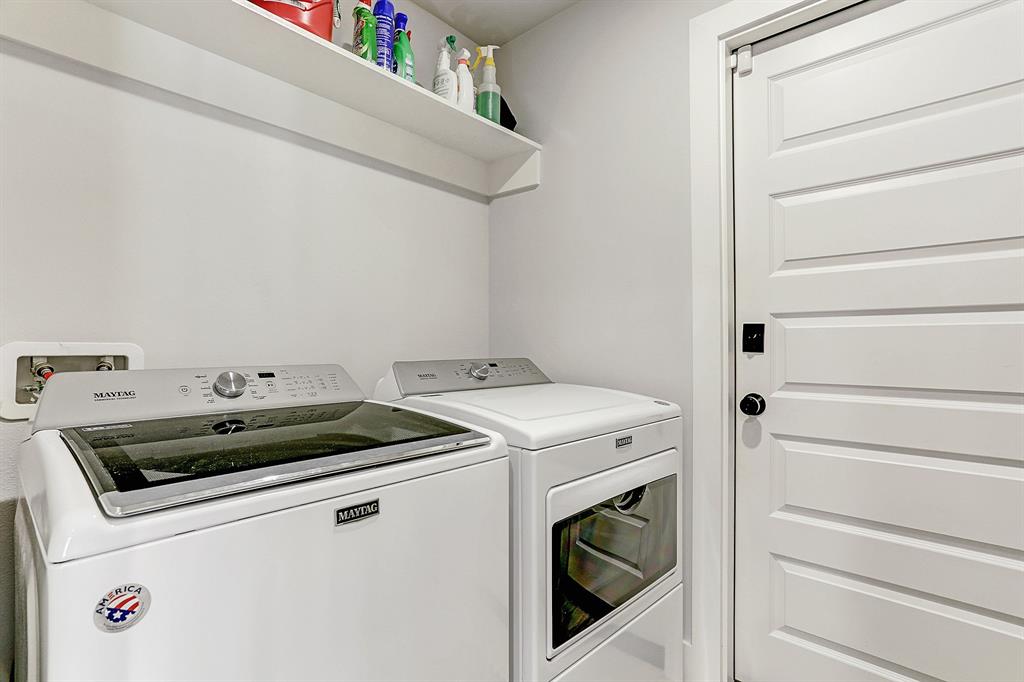 Utility room with built-in shelving above the washer/dryer and additional shelving on opposite wall. Door leads to two-car garage.