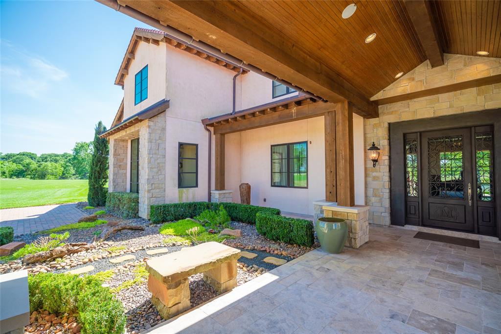 A grand motor court ENTRY to the Main House with its  covered stained wood planked pitched portico ceiling with recessed lighting, hanging chandelier, stone pillars, stone tile flooring, handsome glass/wood front door/flanking windows with decorative iron design, noteworthy light sconces, outdoor speakers - all surrounded by well maintained rock gardens and stone benches.