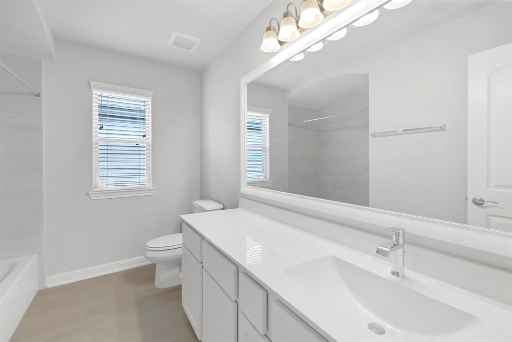 Secondary bath features tile flooring, bath/shower combo with tile surround, white stained wood cabinets, beautiful light countertops, mirror, dark, sleek fixtures and modern finishes.