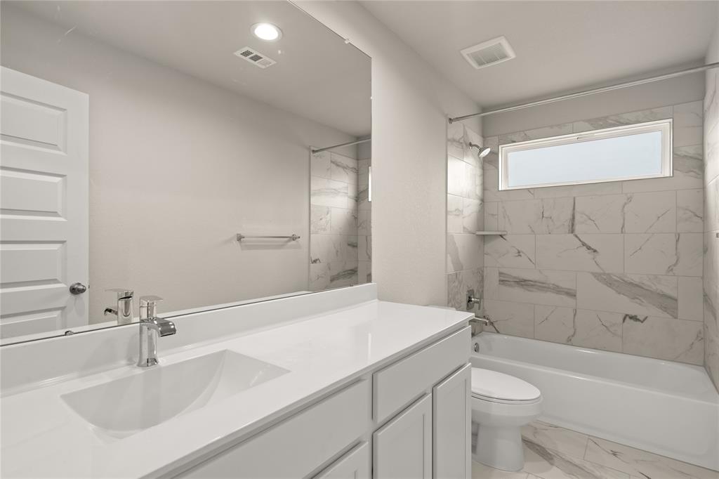 Secondary bath features tile flooring, bath/shower combo with tile surround, white stained wood cabinets, beautiful light countertops, mirror, dark, sleek fixtures and modern finishes.