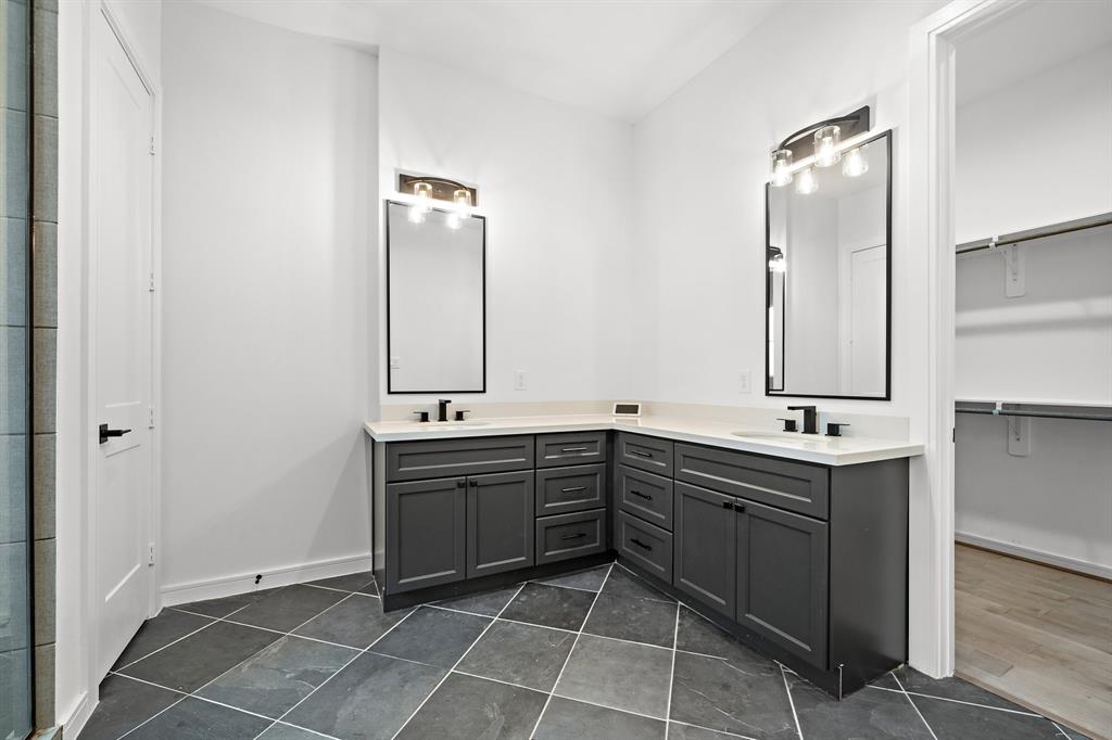 The primary ensuite bath highlights sleek tile flooring, a walk-in closet, and a vanity area with ample built-in storage, double sinks, and tall vanity mirrors with elegant wall sconce light fixtures.