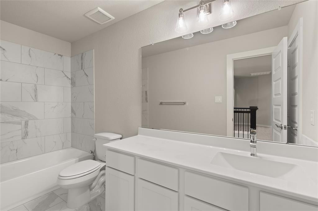 Secondary bath features tile flooring, bath/shower combo with tile surround, white stained wood cabinets, beautiful light countertops, mirror, dark, sleek fixtures and modern finishes.