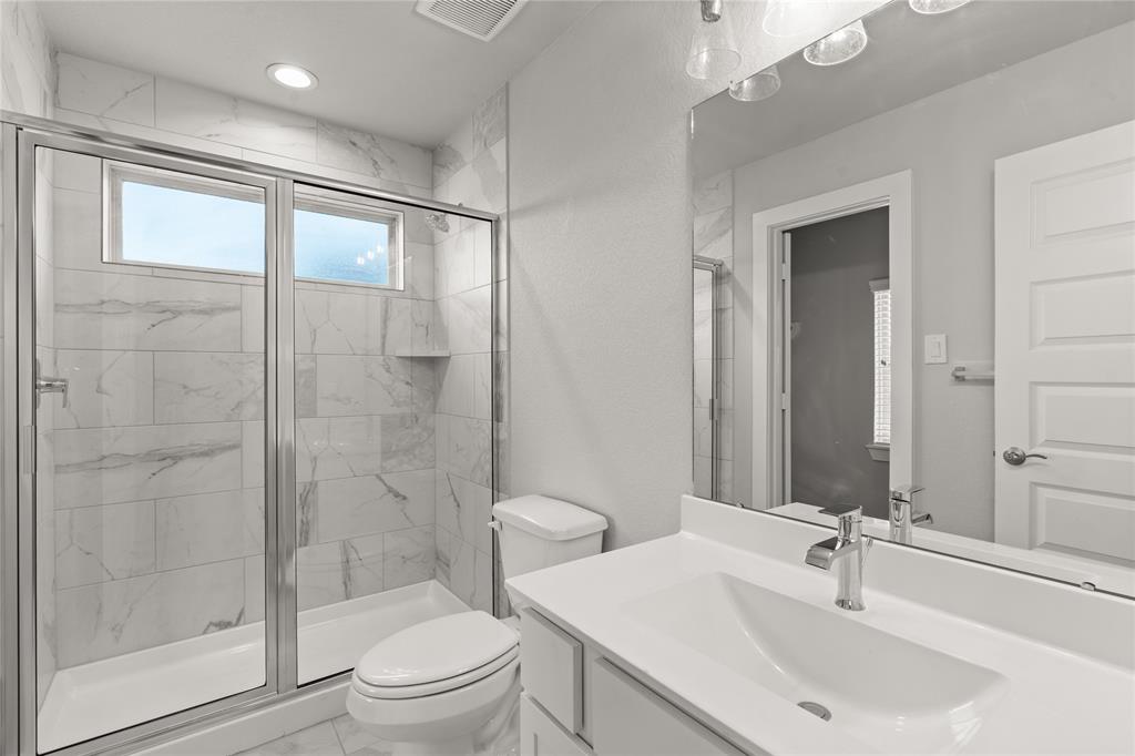 This private bath features tile flooring, shower with tile surround, cool wood cabinets, beautiful light countertops, mirror, dark, sleek fixtures and modern finishes.