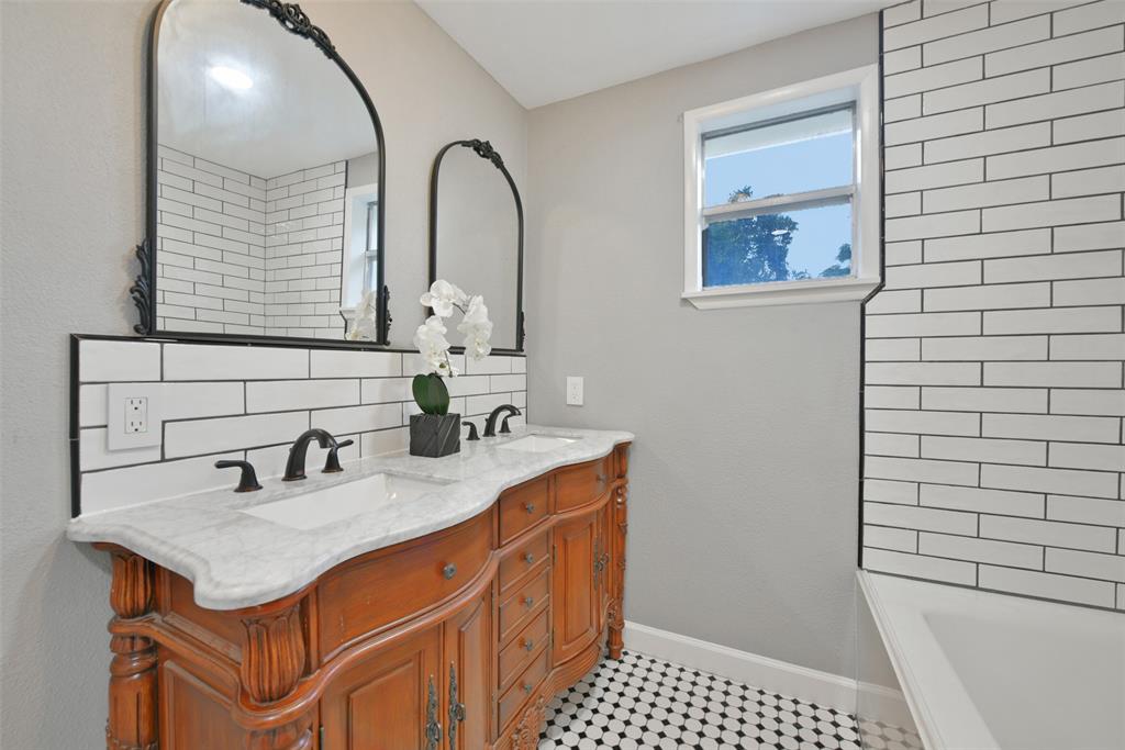 This upstairs bathroom services the bedrooms and it boasts an ornate dresser-style vanity with marble countertops and dual rectangular basin sinks, antique framed mirrors, black and white tile floors, and a combination bathtub and shower with a subway tile surround.