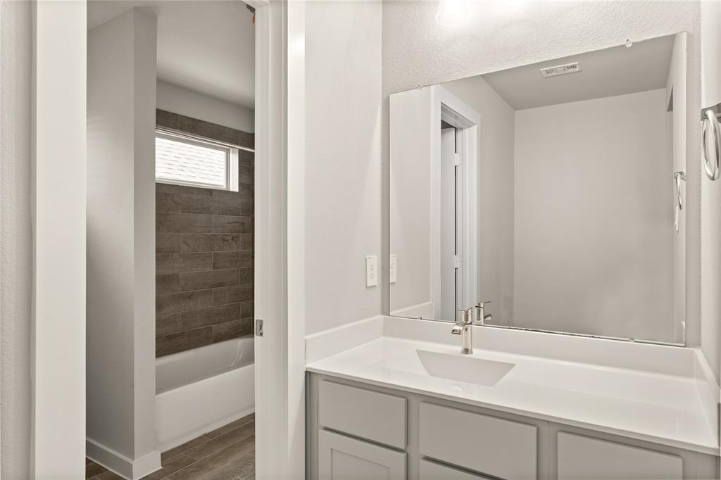 The Jack and Jill bath features tile flooring, bath/shower combo with tile surround, light stained wood cabinets, beautiful light countertops, mirror, dark, sleek fixtures and modern finishes.