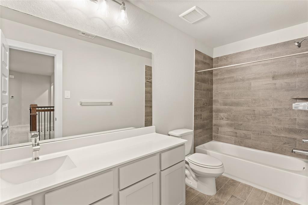 Secondary bath features tile flooring, bath/shower combo with tile surround, light stained wood cabinets, beautiful light countertops, mirror, dark, sleek fixtures and modern finishes.