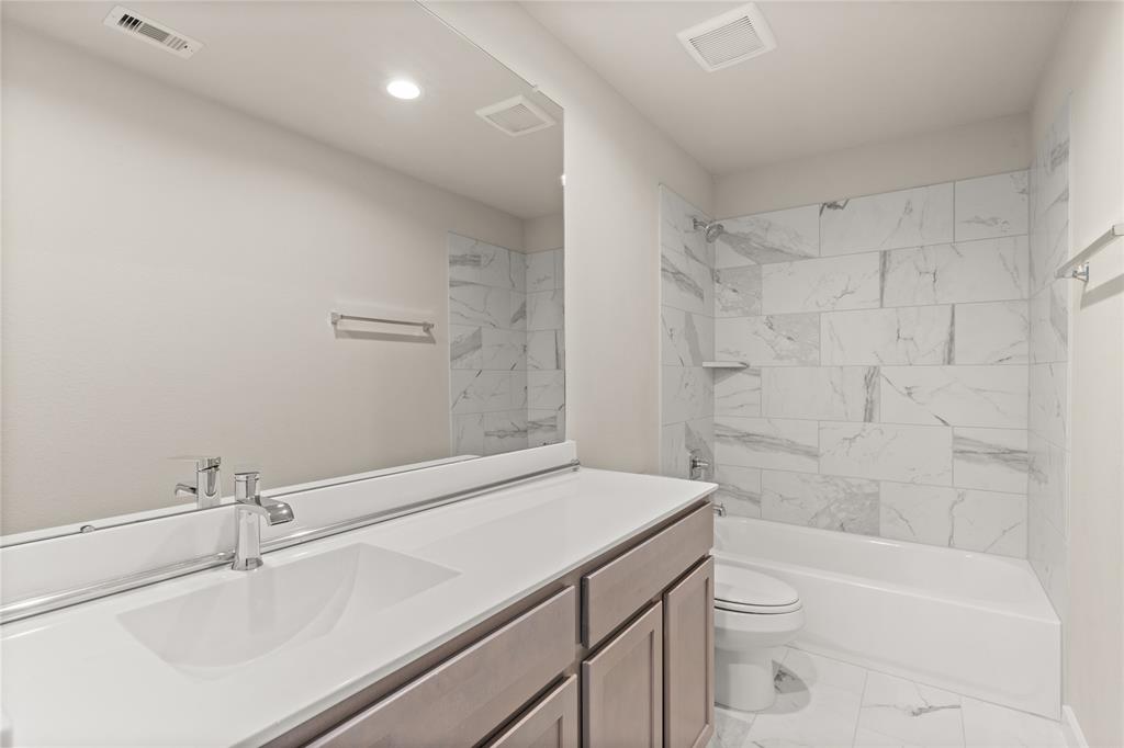 Secondary bath features tile flooring, bath/shower combo with tile surround, light stained wood cabinets, beautiful light countertops, mirror, dark, sleek fixtures and modern finishes.