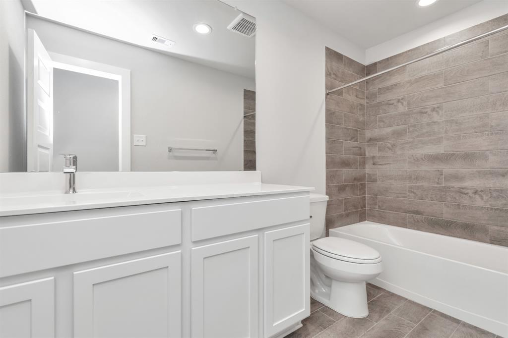 The secondary bathroom boasts tiled floors, a bath/shower combination enclosed in tiled surround, complemented by light-toned wooden cabinets, and adorned with a stunning light-colored countertop.