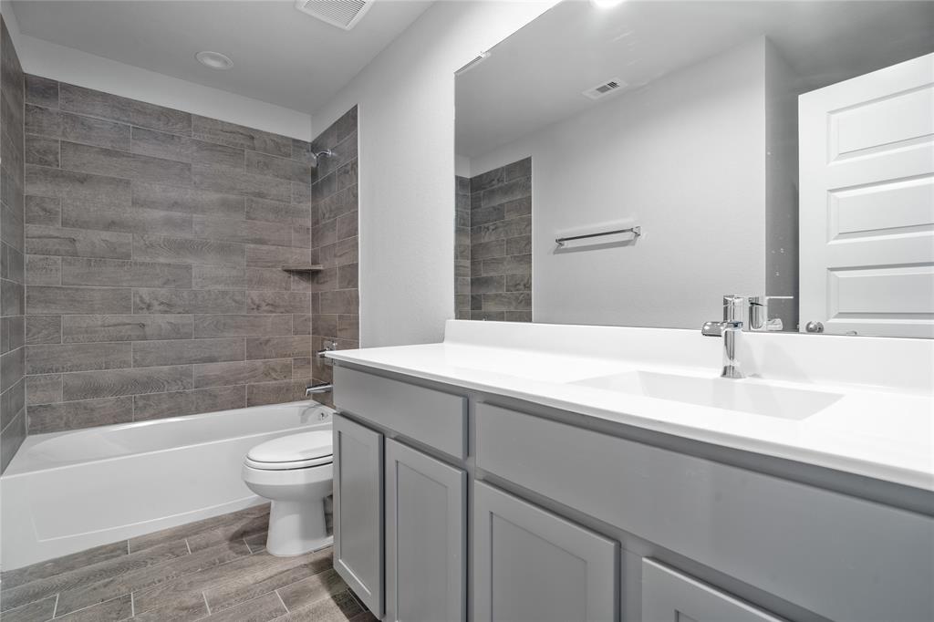 Secondary bath features tile flooring, bath/shower combo with tile surround, stained wood cabinets, beautiful light countertops, mirror, dark, sleek fixtures and modern finishes.