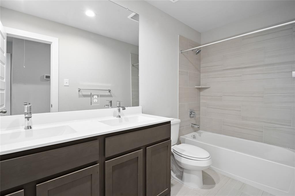 Secondary bath features tile flooring, bath/shower combo with tile surround, dark stained wood cabinets, beautiful light countertops, mirror, dark, sleek fixtures and modern finishes.