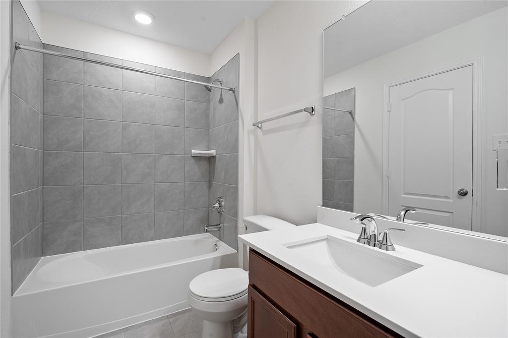 Secondary bath features tile flooring, bath/shower combo with tile surround, stained wood cabinets, beautiful light countertops, mirror, dark, sleek fixtures and modern finishes.