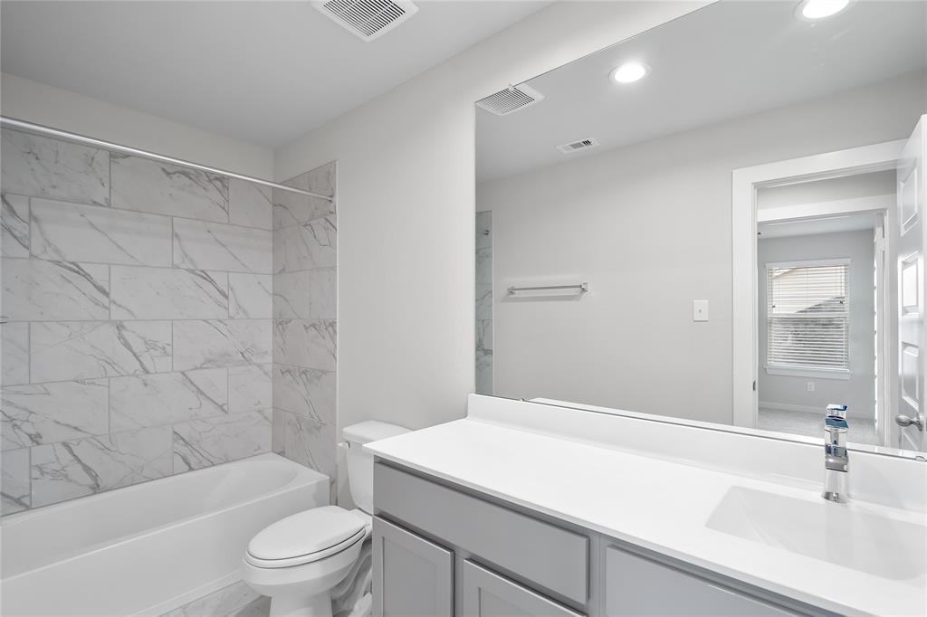 Secondary bath features tile flooring, bath/shower combo with tile surround, white stained wood cabinets, beautiful light countertops, mirror, dark, sleek fixtures and modern finishes.