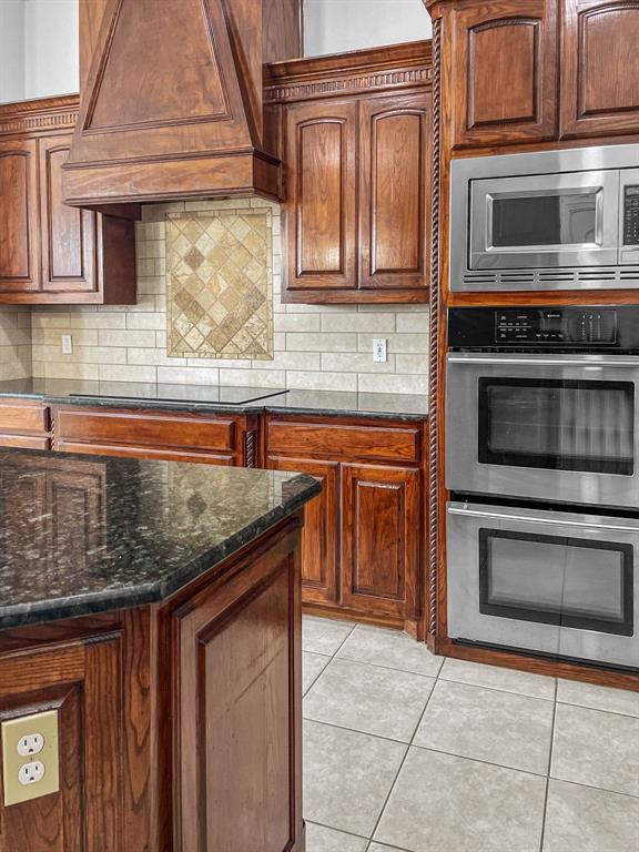 Kitchen featuring backsplash, light tile flooring, stainless steel microwave, double oven, custom exhaust hood, and black electric stovetop