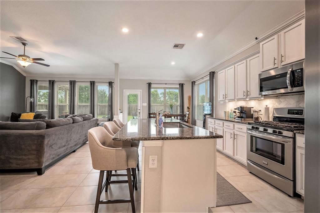 Modern kitchen with white cabinets and stainless steel appliances, opening to a living area with a large gray sofa and windows.