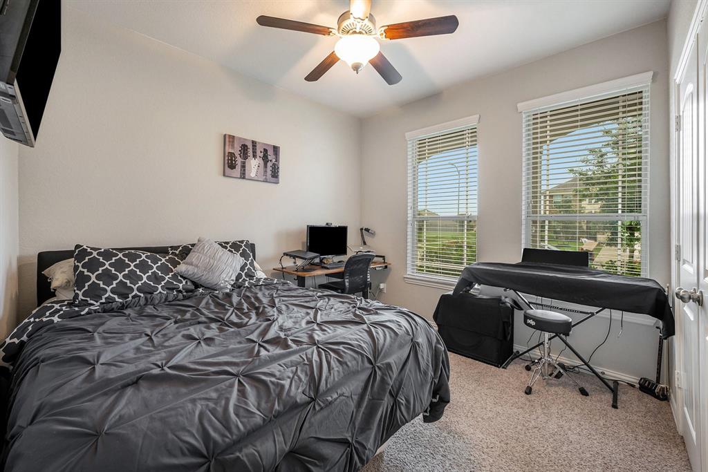 A downstairs bedroom with a bed, desk, computer, electronic keyboard, and two windows with blinds, featuring a monochromatic color scheme and a ceiling fan.