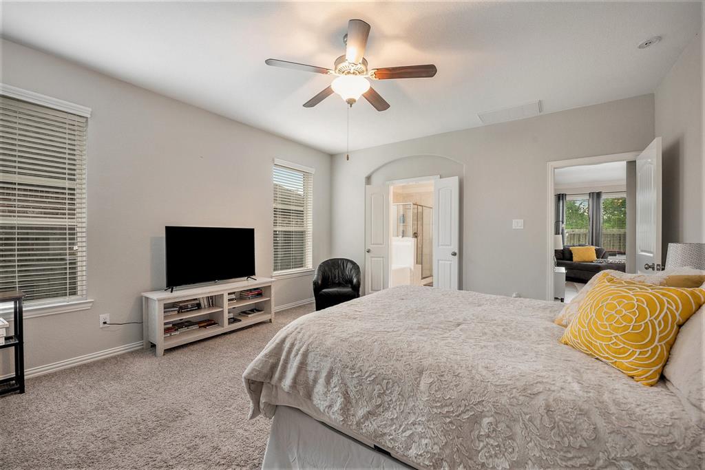 A well-lit, tidy primary bedroom with a large bed, ceiling fan, television, windows with blinds, and an adjoining bathroom visible through an open door.