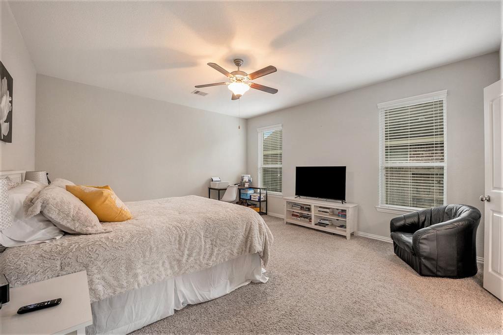 A neat primary bedroom with a large bed, white bedding, a TV stand, a black armchair, two windows, and a ceiling fan.