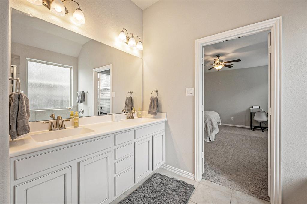 A tidy primary bathroom with double vanity sinks, large mirror, frosted window, and a view into an adjoining room has a ceiling fan.