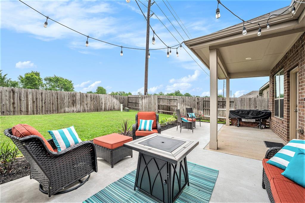 A cozy backyard patio with wicker furniture, colorful cushions, a fire pit, string lights, and a well-maintained lawn enclosed by a wooden fence.