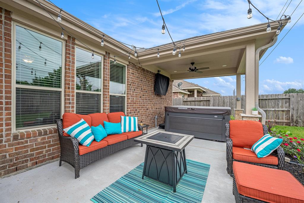 A cozy covered patio with wicker furniture, colorful cushions, string lights, a hot tub, and a fan, adjacent to a brick house.