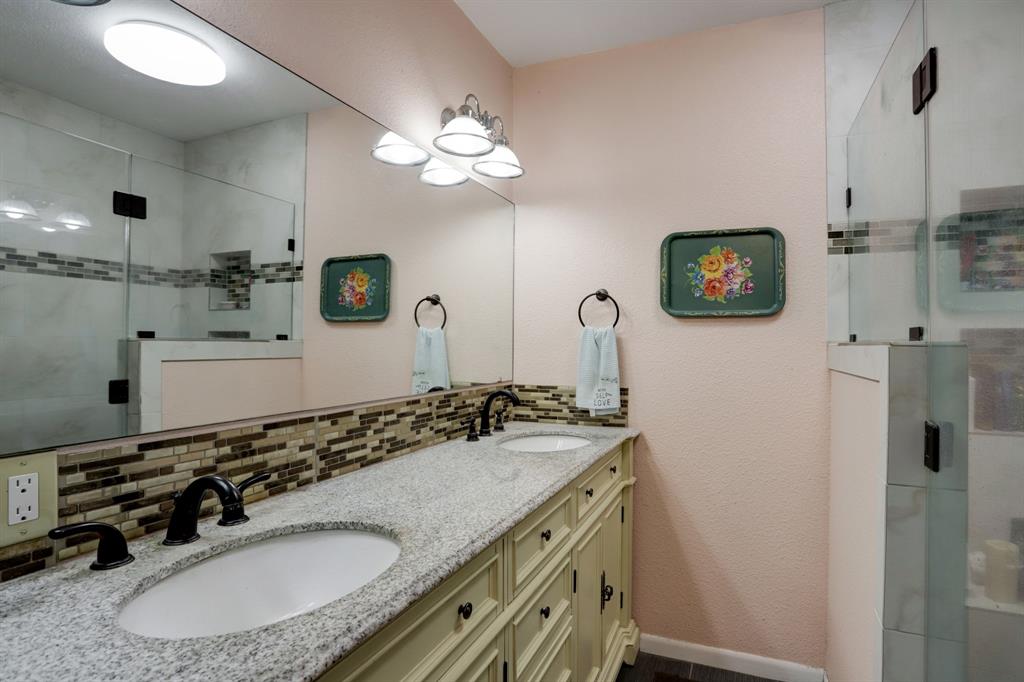 A bathroom with dual sinks, a large mirror, tiled backsplash and a glass shower enclosure.