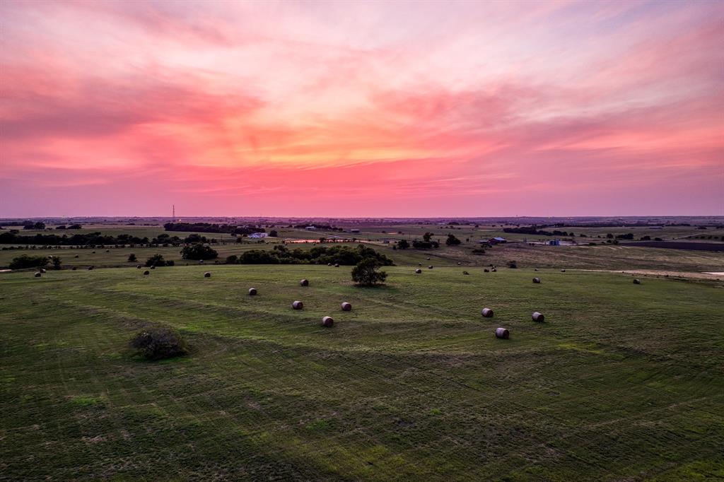 The setting sun casts a warm, golden glow over these Texas rolling hills and verdant fields, highlighting the natural beauty of the landscape. Bales of hay dot the lush green pastures, their shadows stretching long and soft against the earth as the light begins to fade.