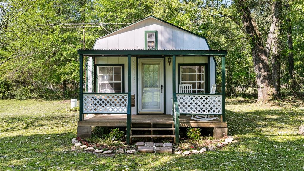 Charming small cottage with green trim, front porch, white railing, surrounded by lush trees, exuding a peaceful, rustic vibe in a serene setting.