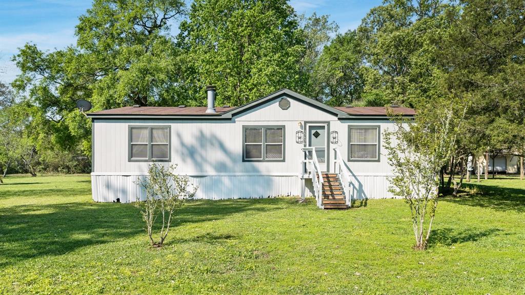 Exterior front main home: Single-story manufactured house with white siding, dark trim, steps leading to a central door, surrounded by trees and green lawn.