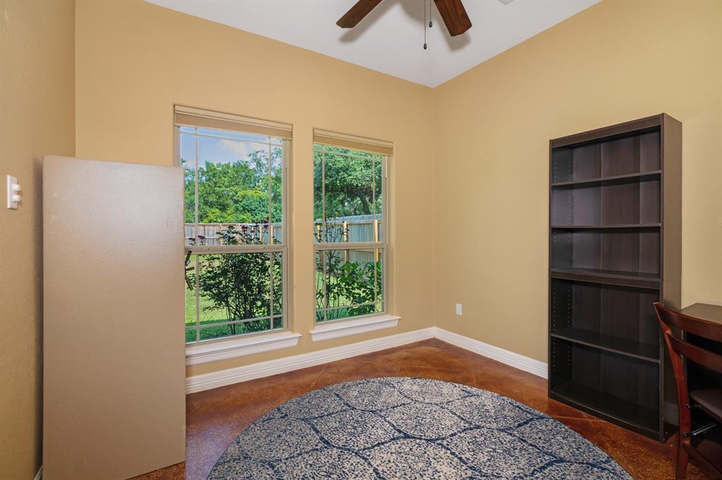 Bright, cozy home office with large windows, wooden ceiling fan, bookshelf, and a large gun safe, overlooking backyard.