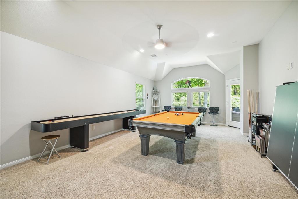 Spacious gameroom with beige carpet, vaulted ceiling, pool table, shuffleboard, bar stools, window, door to outside, and a ceiling fan.