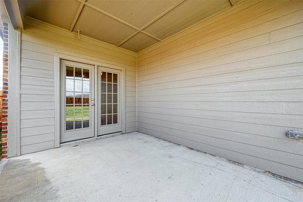 A covered patio with a concrete floor and double doors leading to a grassy backyard, enclosed by a wooden fence.