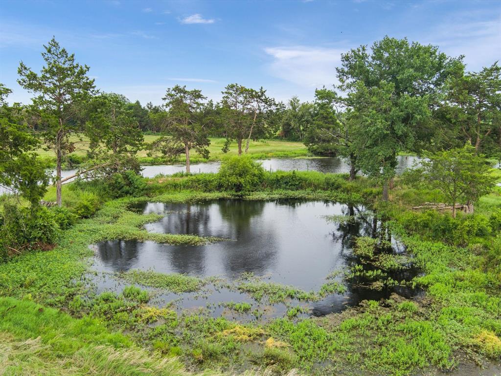 Aerial view of a serene rural landscape with a pond, green fields, trees, and round hay bales under a cloudy sky.