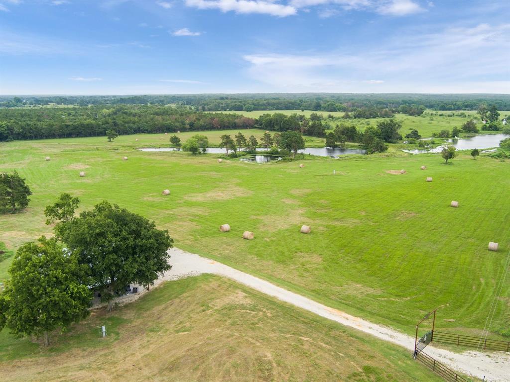 Aerial view of a rural landscape with hay bales, a dirt road, trees, ponds, and vast green fields under a cloudy sky.