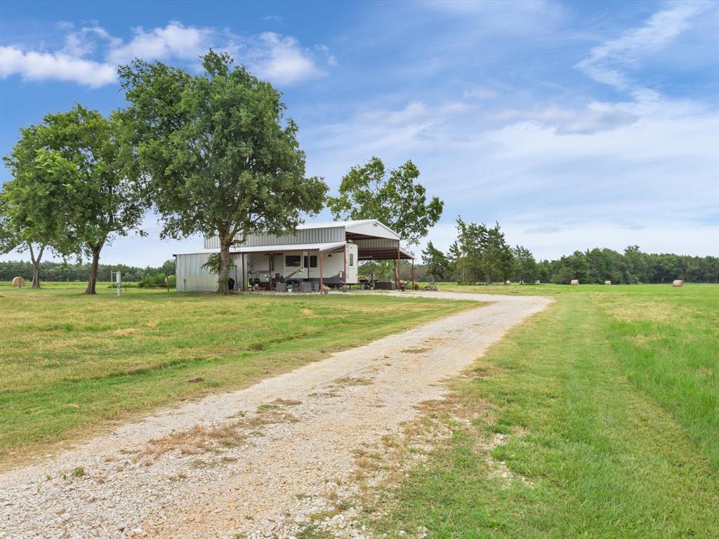 Rural landscape with a gravel driveway leading to a custom metal RV barn, flanked by trees and a field with hay bales under a cloudy sky.