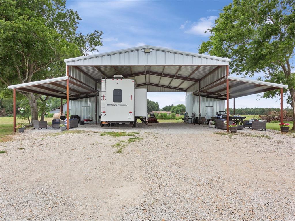 A largecustom metal barn sheltering an RV with outdoor furniture and grills, set in a rural area with trees and a gravel ground.