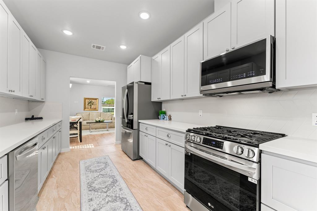 This photo features a modern kitchen with sleek stainless steel appliances, white cabinetry, and ample counter space. It has a bright and open layout with recessed lighting and a view into a cozy living area.