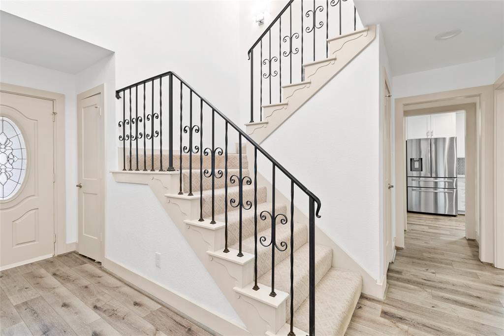 Enter your bright and airy foyer, where natural light flows through the space, highlighting the wood-look flooring and the decorative front door with leaded glass details. The staircase, featuring classic wrought-iron balusters and gently patterned carpet, adds warmth and elegance. Offering easy access to the kitchen and main living areas, this entryway seamlessly blends style with functionality.