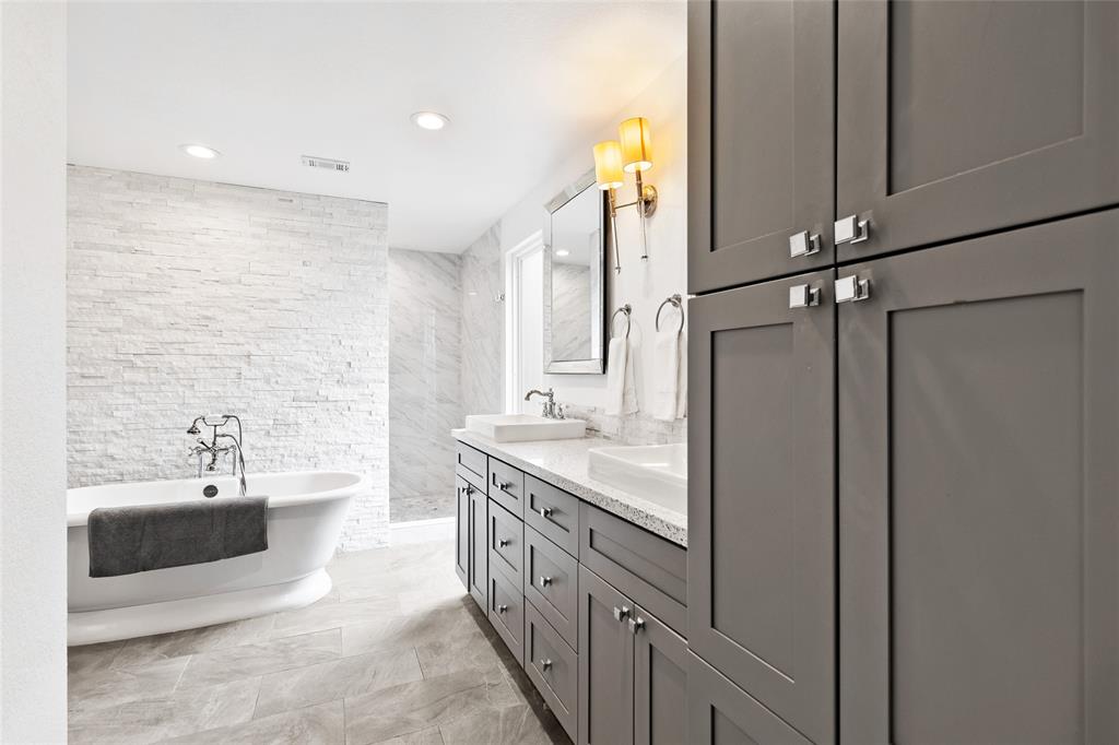 Step into your serene primary bathroom, featuring a freestanding soaking tub against a textured stone accent wall. The walk-in shower, with a modern rainfall showerhead, offers a refreshing escape. A double vanity with quartz countertops and sleek fixtures combines style and functionality. Natural light highlights the luxurious finishes, enhancing the calming atmosphere of your retreat.