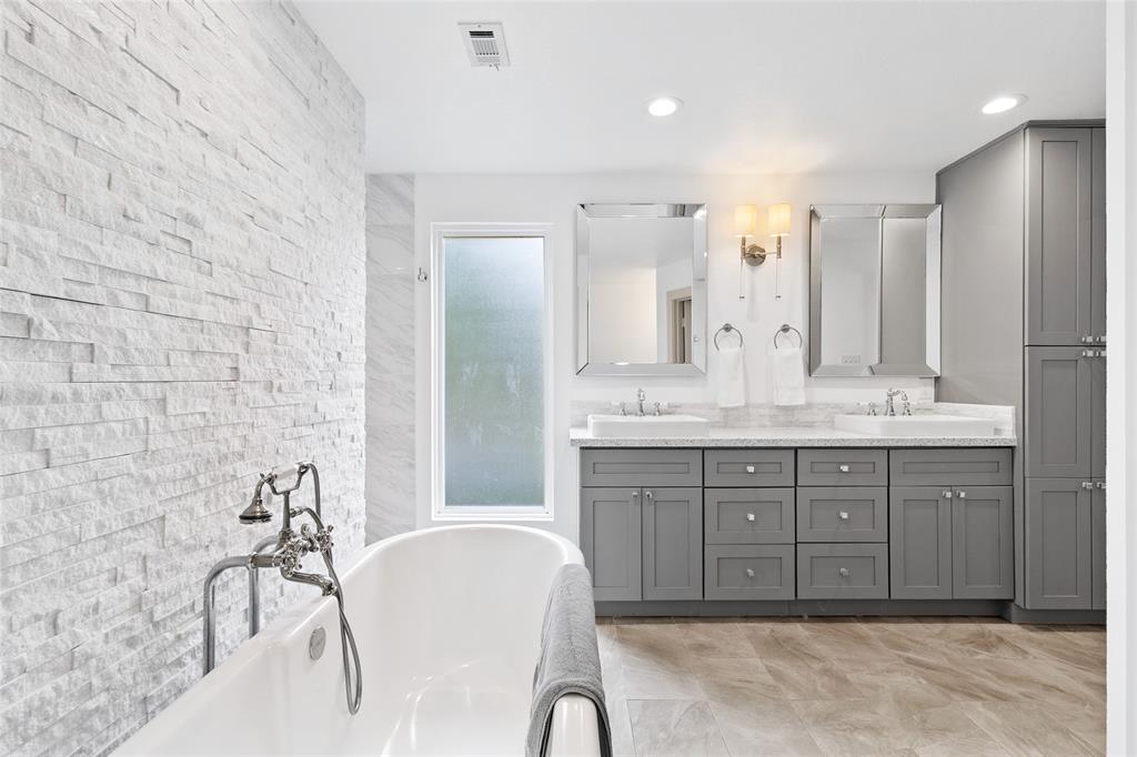 Take another look at your goregous primary bathroom, featuring a modern double-sink vanity with sleek grey cabinetry and ample storage. A frosted window allows soft natural light while ensuring privacy, and large-format tile flooring adds sophistication. The freestanding soaking tub, paired with classic chrome fixtures and set against a textured stone accent wall, creates a tranquil, spa-like retreat.