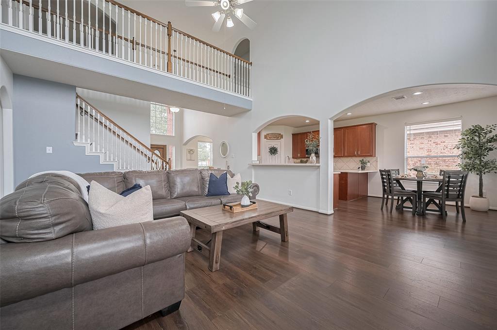 Step into a bright and inviting living room featuring soaring ceilings, expansive windows, and rich wood flooring. The open-concept layout connects seamlessly with the dining area and kitchen, while the fireplace adds a cozy focal point.