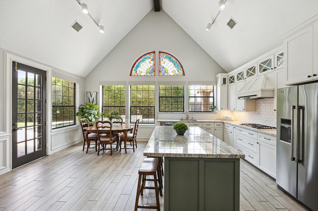 This spacious kitchen features a high vaulted ceiling and large windows that flood the area with natural light. It includes a central island with granite countertops, modern stainless steel appliances, and ample white cabinetry. The dining area offers a charming view outside, enhanced by a decorative stained glass window.