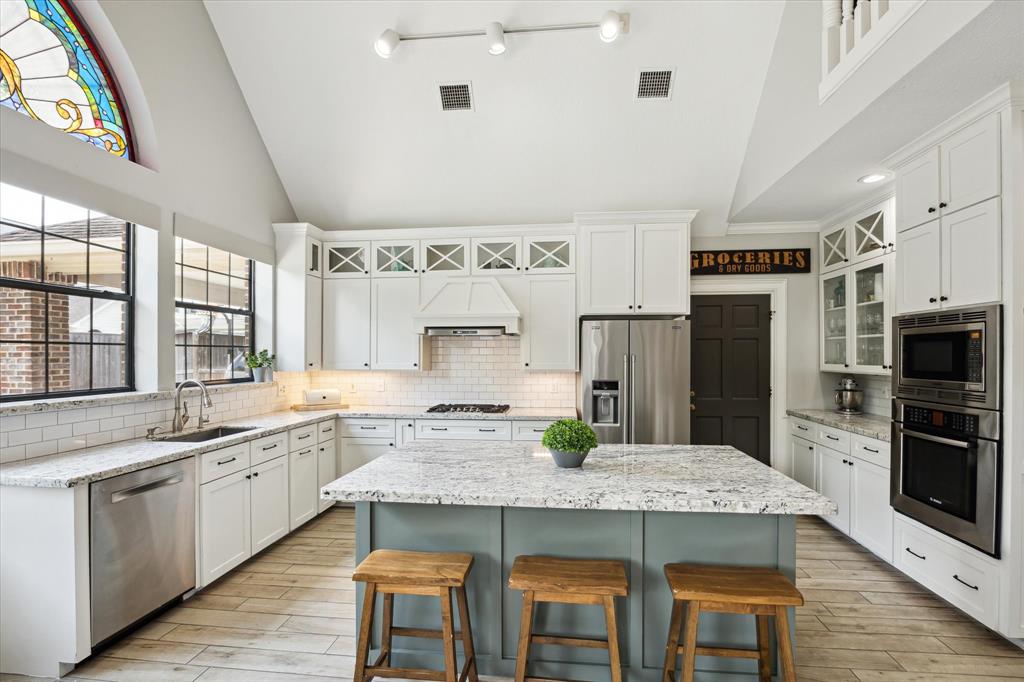 Stunning, light-filled kitchen with soaring vaulted ceilings, custom white cabinetry, granite countertops, and subway tile backsplash. Features a large center island with seating, a gas cooktop (converted from electric), and stainless steel appliances.  Enjoy a beautiful view of the backyard and pool, while the stained-glass transom adds a unique architectural touch.