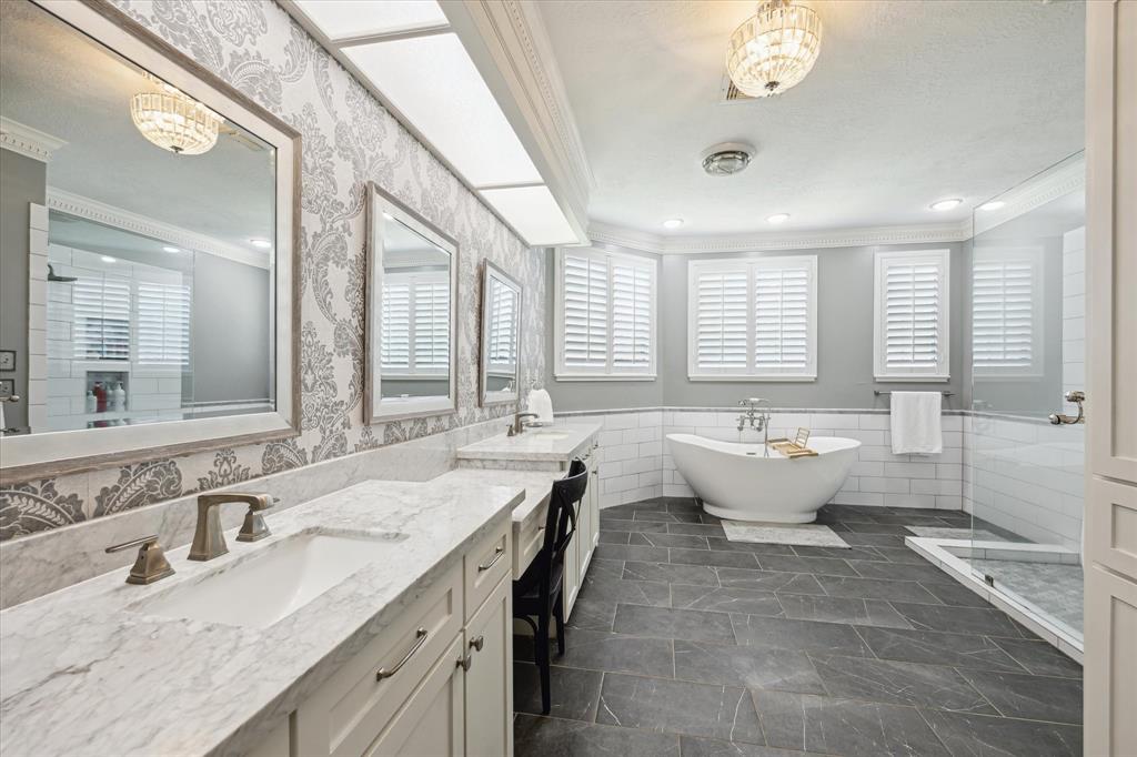 Luxuriously updated primary bathroom featuring a freestanding soaking tub, oversized frameless glass shower, dual marble vanities, and a built-in makeup area. Elegant wallpaper accents, plantation shutters, and rich tile flooring complete this spa-like retreat