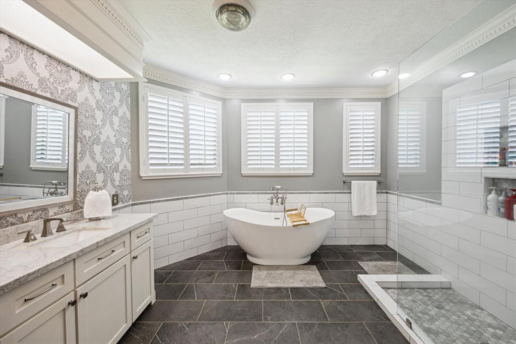 This elegant bathroom features a freestanding tub, glass-enclosed shower, and dual-sink vanity. It boasts modern fixtures, white cabinetry, and stylish tile flooring, with ample natural light from large windows with shutters.