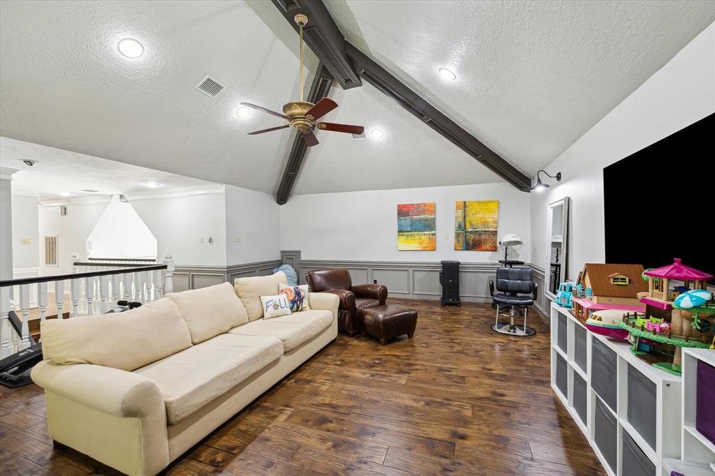 This spacious living area features vaulted ceilings with exposed beams, hardwood floors, and a ceiling fan. It's well-lit with recessed lighting and offers a comfortable seating arrangement. The room is accented with modern artwork and has space for entertainment or play areas.