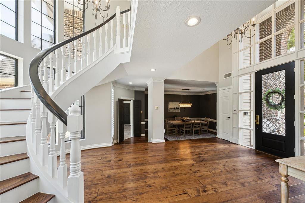 This photo showcases a spacious, elegant entryway with a grand, curved staircase, hardwood floors, and high ceilings. Natural light streams through large windows, and there's a view into a formal dining room. The front door features decorative glass, adding charm and sophistication.