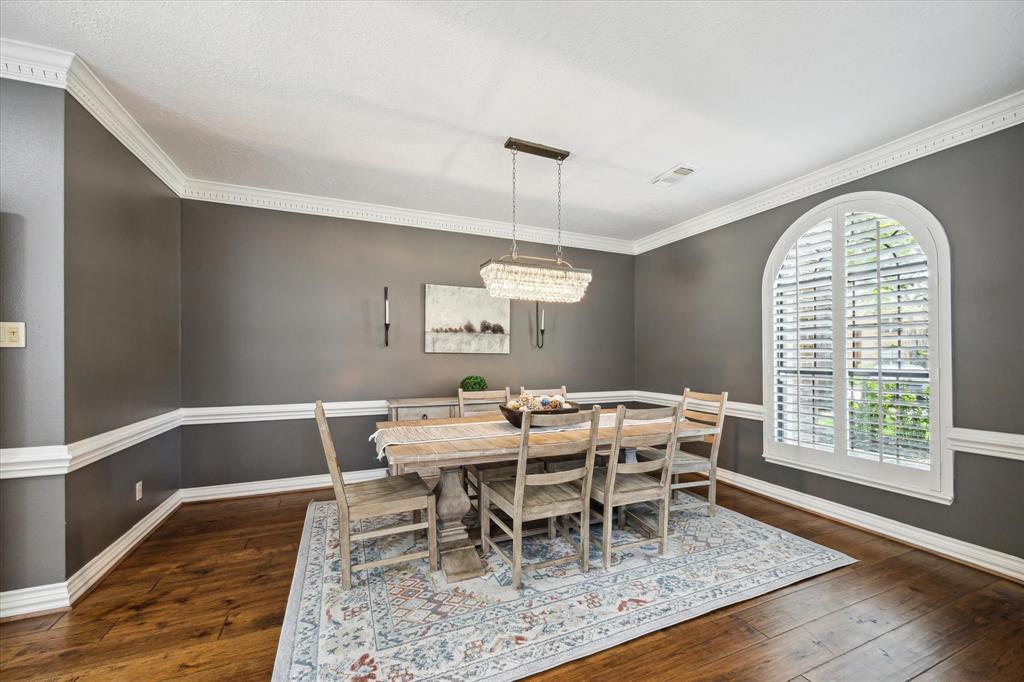 This dining room features elegant dark gray walls with white trim and crown molding. It has a large arched window with shutters, a stylish chandelier, and a wooden table set on a patterned area rug. The hardwood floors add warmth and sophistication.