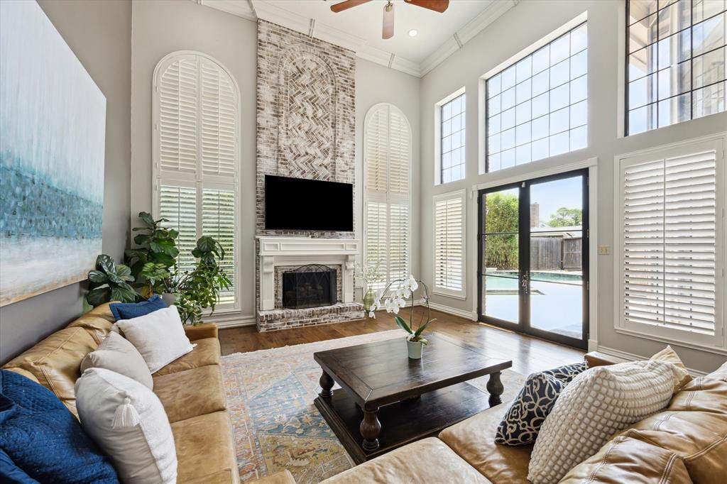 Spacious formal living room with high ceilings and abundant natural light. Features a cozy fireplace, tall windows with shutters, and a view of the backyard pool. Elegant decor includes a large sectional sofa and a central coffee table.