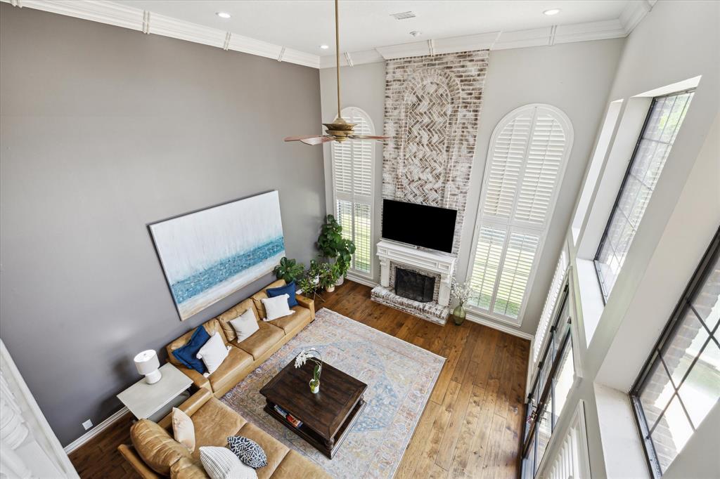 This spacious living room features high ceilings and large windows, allowing for plenty of natural light. It includes a cozy fireplace, modern ceiling fan, and elegant wood flooring. The decor highlights a neutral palette with a stylish accent wall and a comfortable seating area.