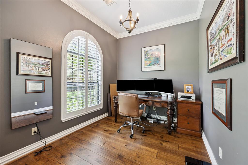 This cozy home office features elegant wood flooring, a classic wood desk with dual monitors, and ample natural light from a large arched window with plantation shutters. The room is enhanced by tasteful wall art and a stylish chandelier, offering a refined and productive workspace.